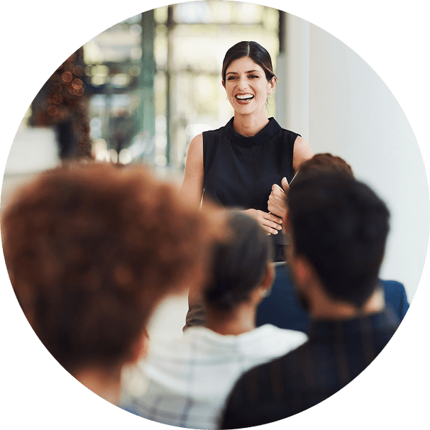 A woman in front of a seated group, smiling. She holds a clipboard