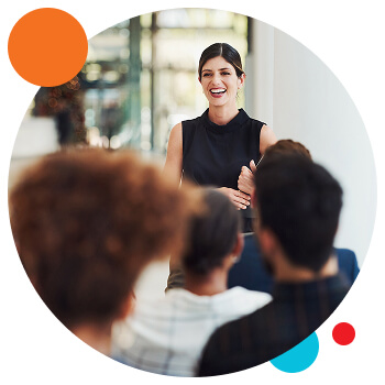 HP circle - wellbeing A woman standing in front of a seated crowd. She is smiling and holding a clipboard.
